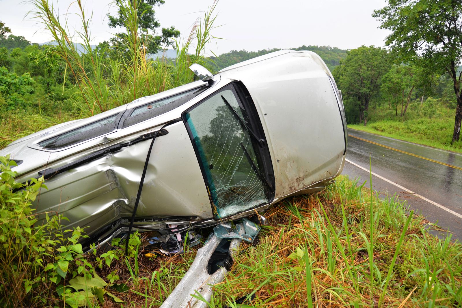 A silver SUV rolled onto its side in the grass beside a wet road after a single-vehicle rollover crash
