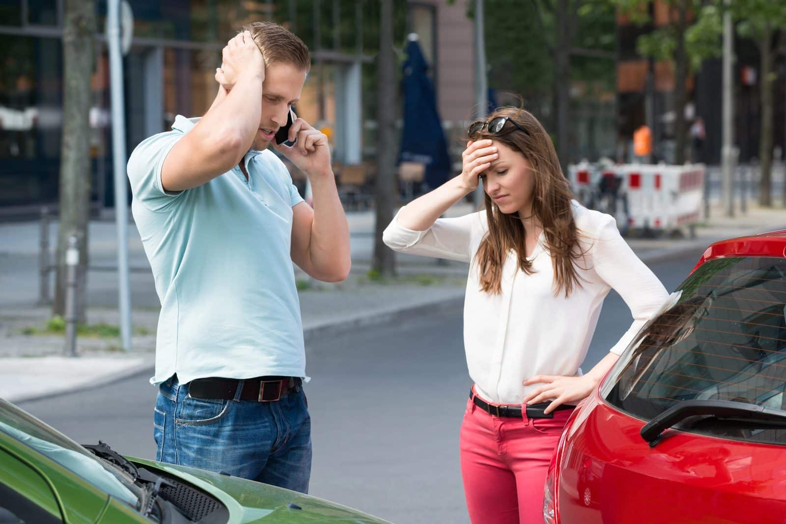 Two stressed drivers beside a damaged car after a collision, one on the phone with an insurer.